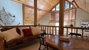 A cozy loft-style room with a wooden ceiling, a sofa with cushions, a dining table, and a view of the outdoors through large windows. - 
Centerport GetAway, Family size cabin. Near restaurants, golf and AU football (Dadeville)