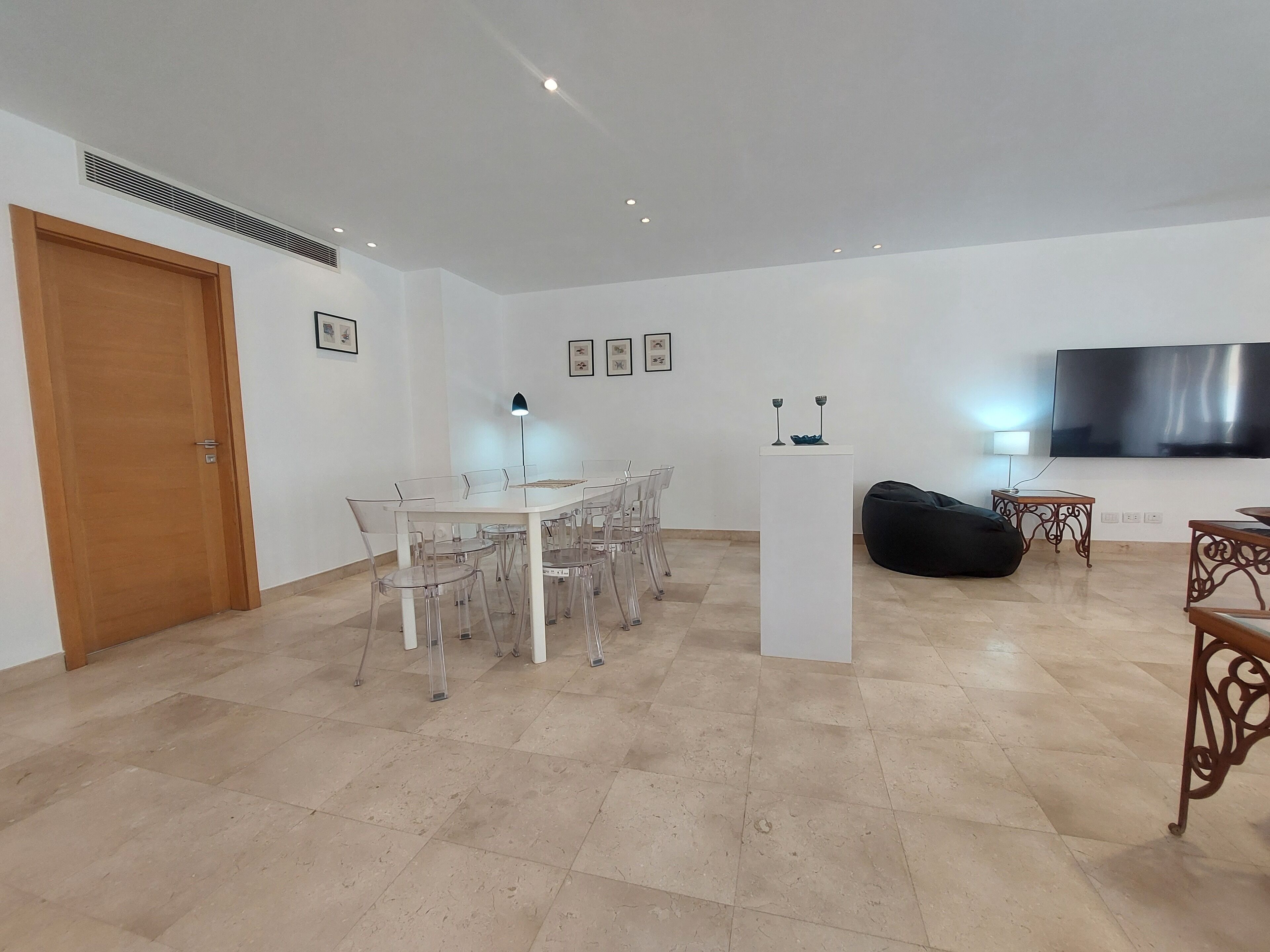 A spacious living area with a dining table, chairs, a black bean bag, a flat-screen TV, and a white pedestal.