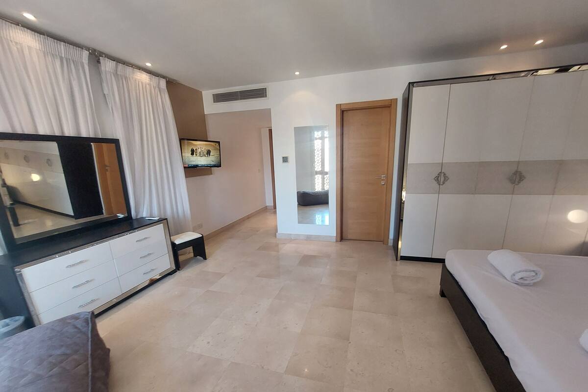 A modern hotel room with a large bed, a TV mounted on the wall, a dresser, and a seating area with a chair and a small table.