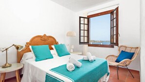 A bedroom with a wooden bed, a view of the sea through an open window, a wicker chair, and a small bedside table with a lamp. - Sunset Tirant, Your seaside retreat (Platges de Fornells)