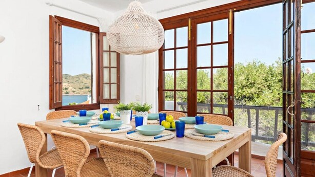A dining area with a wooden table set for four, surrounded by wicker chairs, with a view of greenery and hills through large windows. - Sunset Tirant, Your seaside retreat (Platges de Fornells)