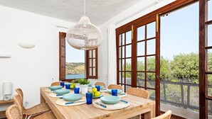A dining area with a wooden table set for six, surrounded by wicker chairs, a hanging light fixture, and large windows with a view of greenery. - Sunset Tirant, Your seaside retreat (Platges de Fornells)
