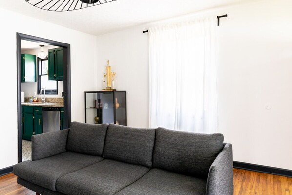 A modern living room with a grey sofa, a black-framed mirror, and a glass cabinet with decorative items. A kitchen with green cabinets is visible through an open doorway.