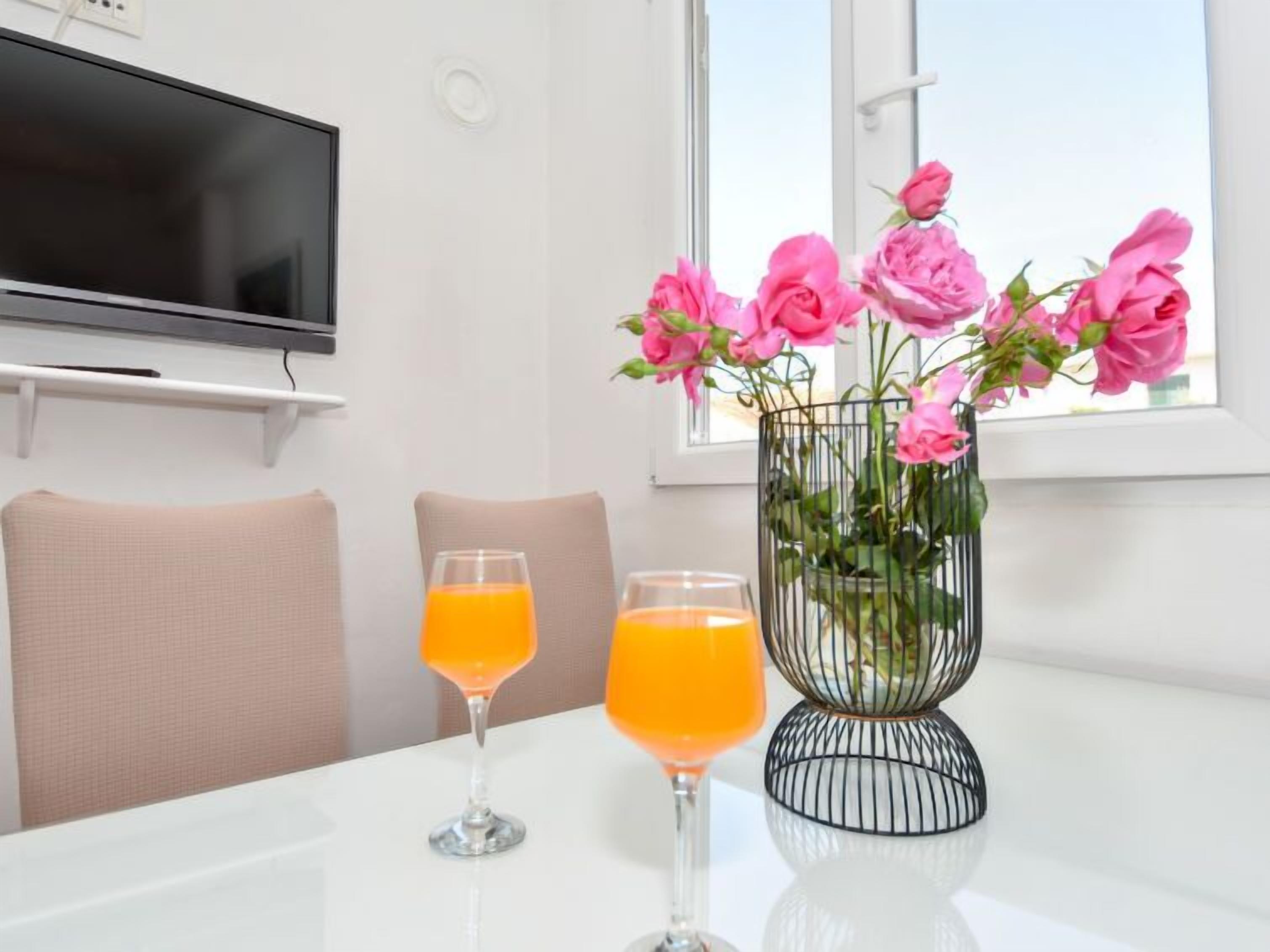 A white table with two orange drinks, a vase with pink flowers, and a TV on the wall.