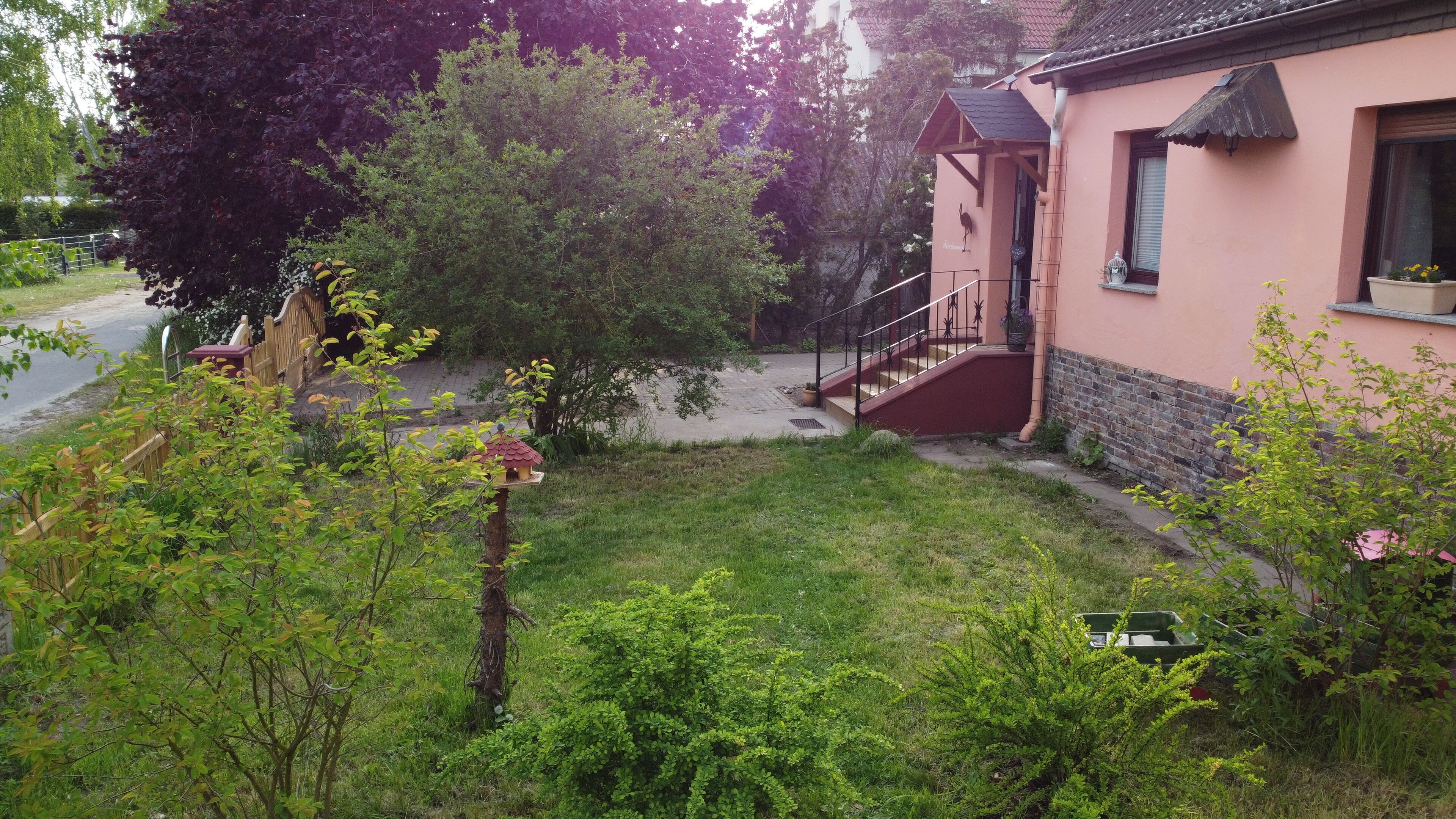Ein Wohngebiet mit einem Haus, das rote Außenwände, eine Holztreppe und einen kleinen Garten mit verschiedenen Pflanzen und einem Vogelhäuschen hat.