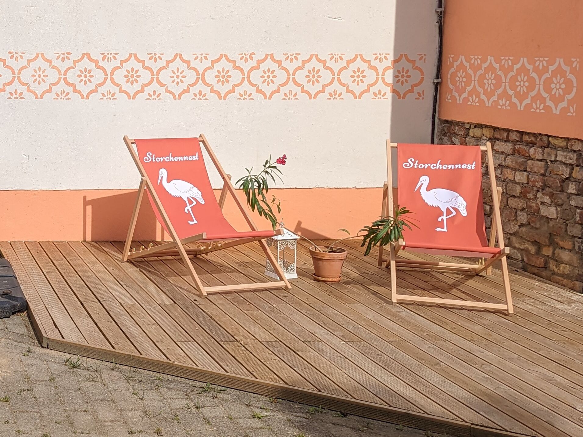 Dos sillas de playa con la marca 'Storchennest' y un logotipo de cigüeña, ubicadas en una terraza de madera con plantas en macetas cerca.