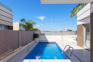 Piscina ao ar livre moderna com céu azul límpido, cercada por um muro de tijolos e palmeiras.