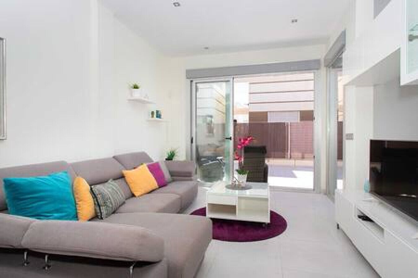 A modern living room with a grey sofa, colorful cushions, a flat-screen TV, and a glass door leading to an outdoor area.