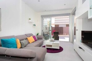 Uma sala de estar moderna com um sofá cinzento, almofadas coloridas, uma televisão de tela plana e uma porta de vidro que dá para uma área externa.