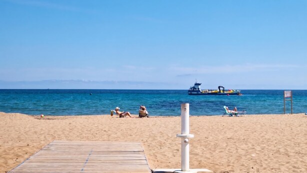 A beach with a wooden walkway leading to the water, a few people relaxing, and a large ship in the distance. - La Gallega - Denia, Costa Blanca (Denia)