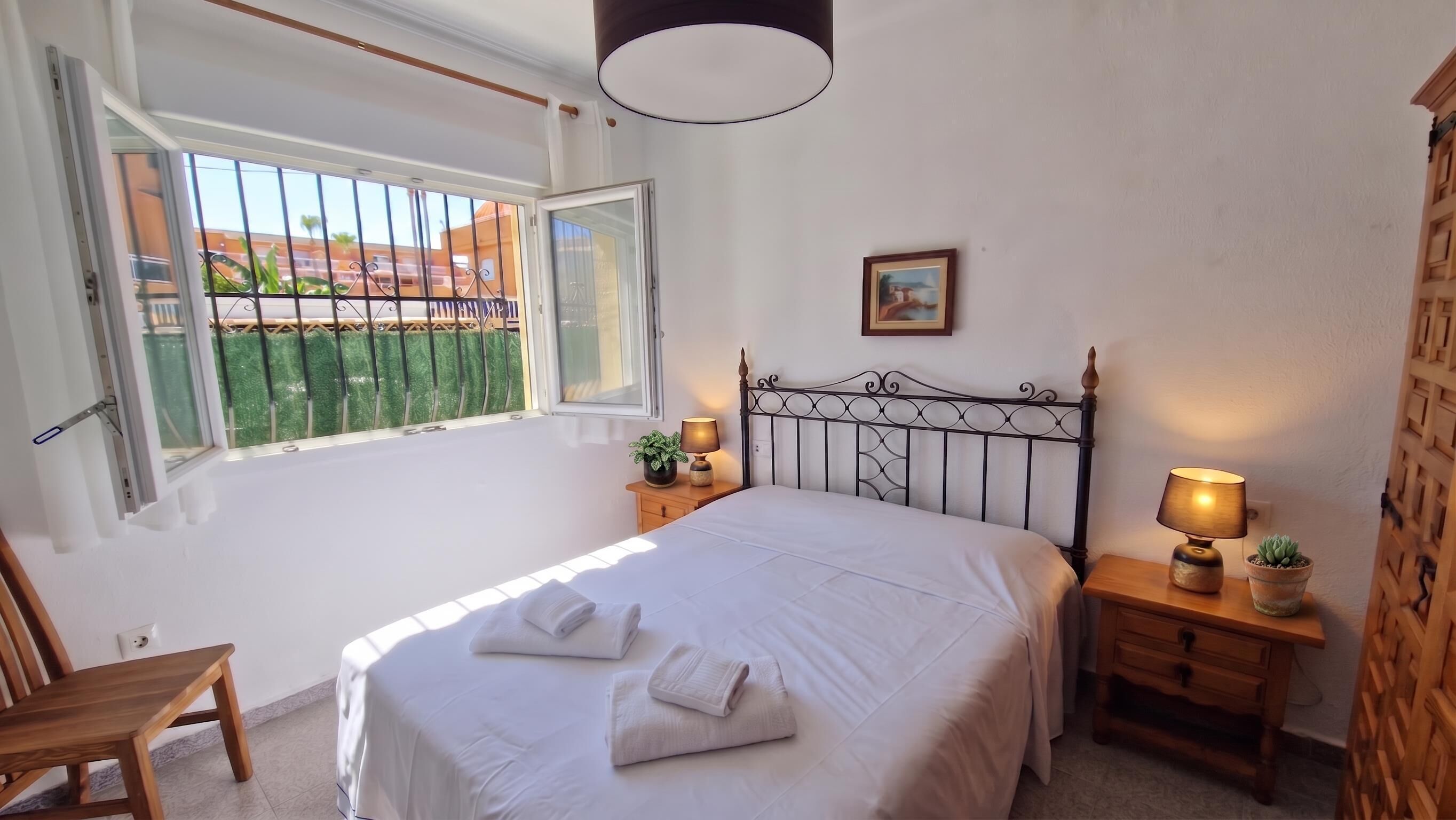 Un dormitorio con cama, dos mesitas de noche con lámparas, una ventana con vistas, una silla y un cuadro colgado en la pared.
