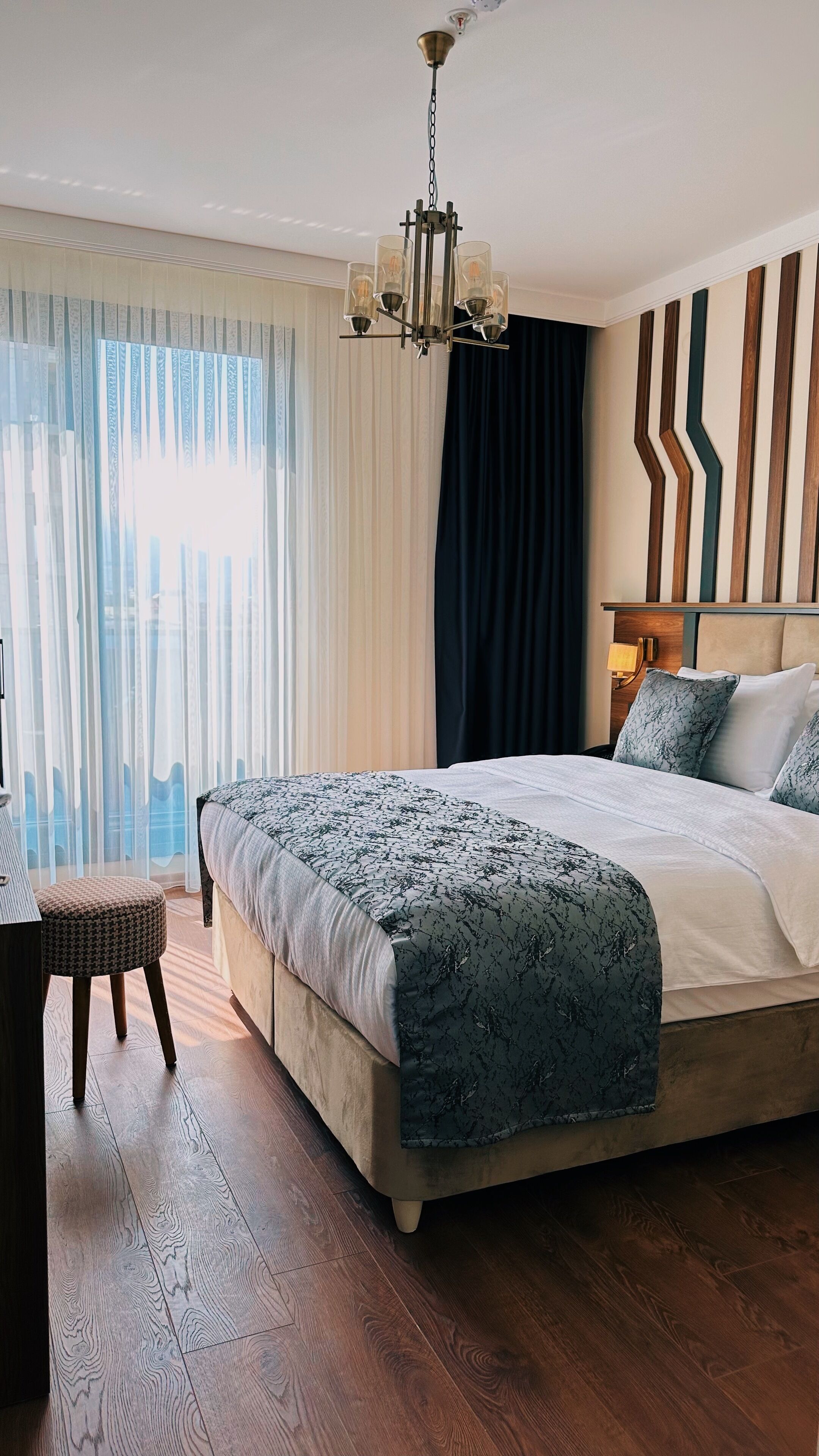Habitación de hotel con una cama grande, suelo de madera, araña y vistas a la ciudad a través de cortinas traslúcidas.