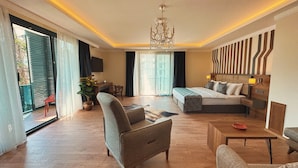 Habitación de hotel con una cama grande, un sofá, un escritorio y un televisor. Se observa una araña, una planta y vistas al exterior.