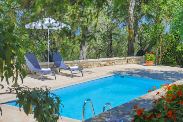 A swimming pool with lounge chairs and an umbrella, surrounded by stone walls and greenery.