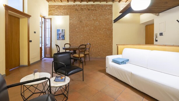 Un cuarto acogedor con pared de ladrillo, sofá blanco, mesa de comedor con sillas y una pequeña mesa redonda con un libro y una vela.