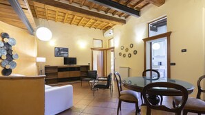 Una sala de estar acogedora con una mesa redonda de comedor, sillas de madera, un sofá blanco y un televisor sobre un mueble de madera.