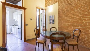 Un comedor con una mesa redonda de vidrio y cuatro sillas, una cocina visible a través de una puerta abierta, y una pared de ladrillo.