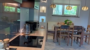 Une cuisine moderne avec une plaque de cuisson à induction noire, une table à manger en bois et des chaises. On y trouve des luminaires suspendus, une fenêtre donnant sur un espace verdoyant et un radiateur placé sous la table.