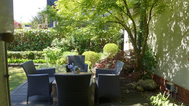 Une terrasse extérieure aménagée avec des meubles en osier et une table, entourée de verdure et avec un bâtiment en arrière-plan.
