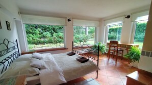 Une chambre avec un grand lit, un bureau et une chaise. Il y a des fenêtres qui donnent sur un paysage verdoyant.
