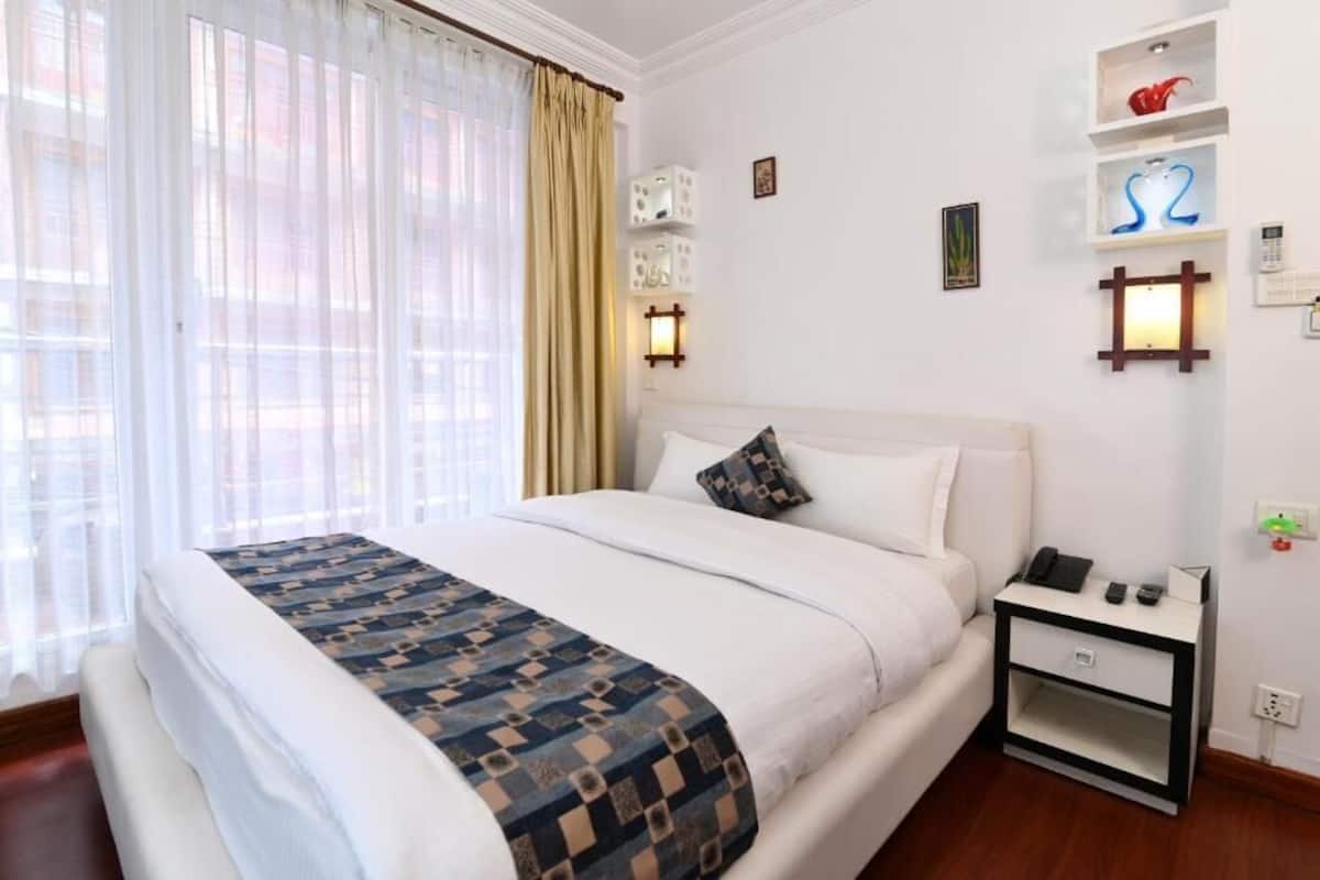 A hotel room with a large bed, a bedside table with a phone and a lamp, and a wall-mounted shelf with decorative items.