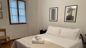 Een netjes opgemaakt bed met een witte deken en kussens, een houten stoel en twee ingelijste kunstwerken aan de muur.