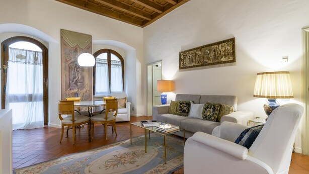 A living room with a sofa, armchair, coffee table, and dining table. There are two lamps, a painting on the wall, and a window with curtains.