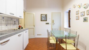 A compact kitchen with white cabinets, a tiled backsplash, a glass dining table with chairs, and a window with decorative plates.