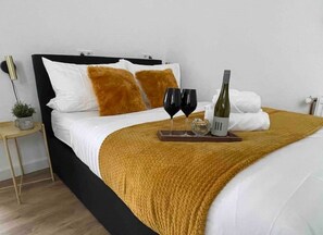 A neatly made bed with a white and mustard-colored blanket, two pillows, a bottle of wine, and two glasses on a tray. - Studio apartment moor-home (Ganderkesee)