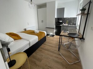 A compact bedroom with a bed, bedside table, and chair. A kitchenette with a microwave, toaster, and small fridge is visible in the background. - Studio apartment moor-home (Ganderkesee)