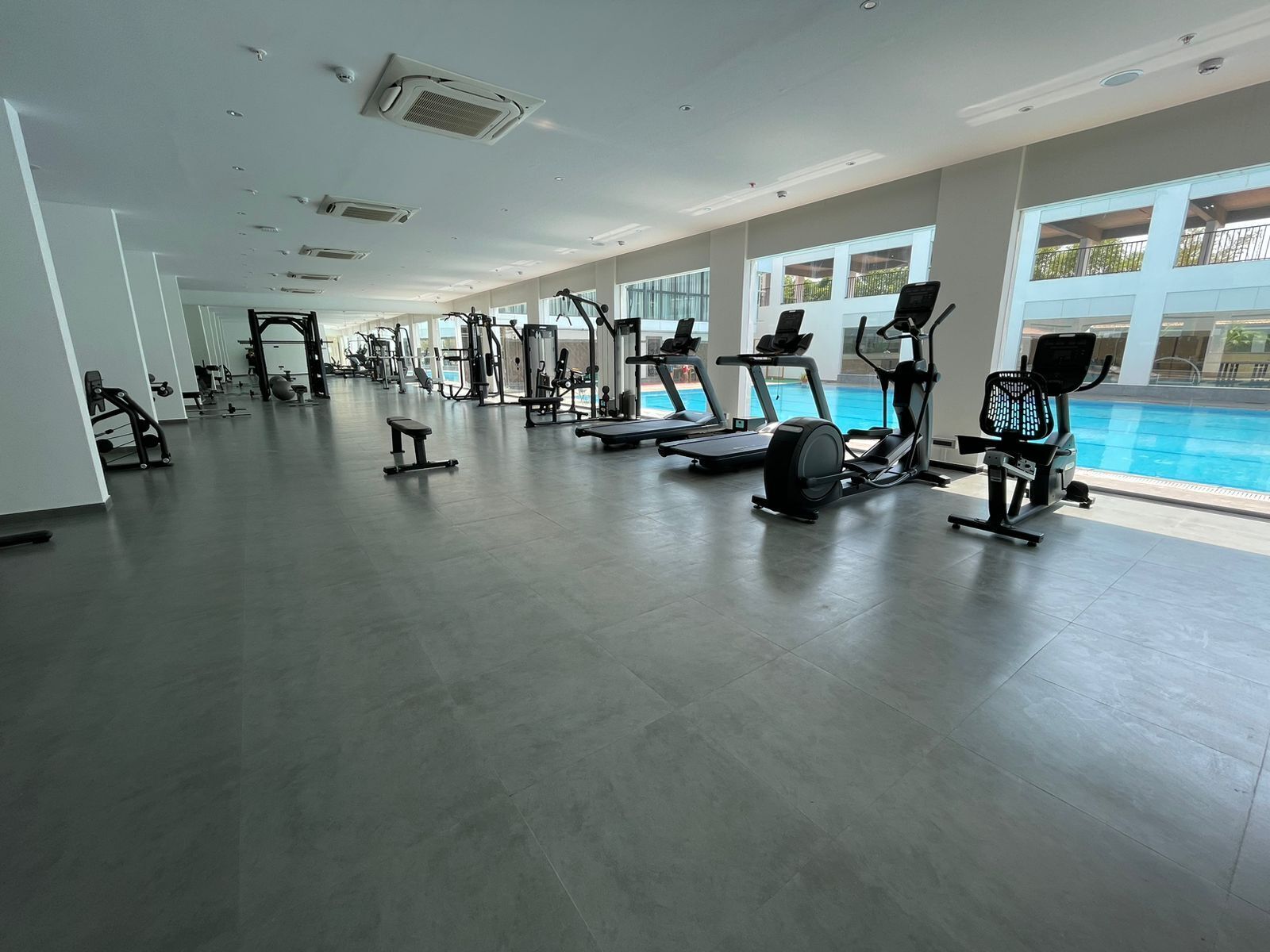 A spacious gym with various exercise equipment, large windows, and a pool area.