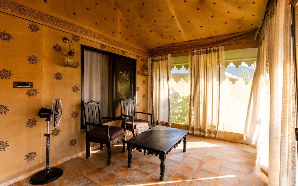 A room with a traditional design, featuring a wooden table and chairs, a standing fan, and a view of the outdoors through large windows with curtains.
