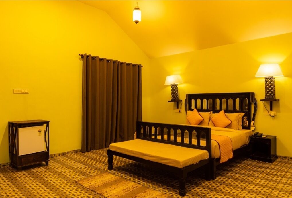 A room with a bed, two bedside lamps, a nightstand, a small table, and a window with curtains.
