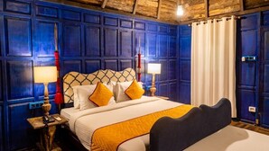 Habitación de hotel con techo de madera, una cama con cabecera estampada, dos mesitas de noche con lámparas y una ventana con cortinas.