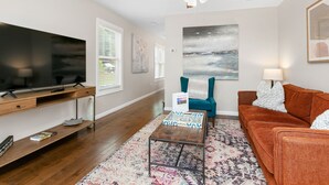 A modern living room with a flat-screen TV, a ceiling fan, a teal armchair, a brown sofa, and a patterned rug.