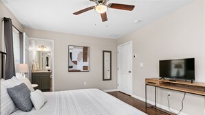 A bedroom with a large bed, a ceiling fan, a TV on a wooden console, and a bathroom with a mirror and sink.