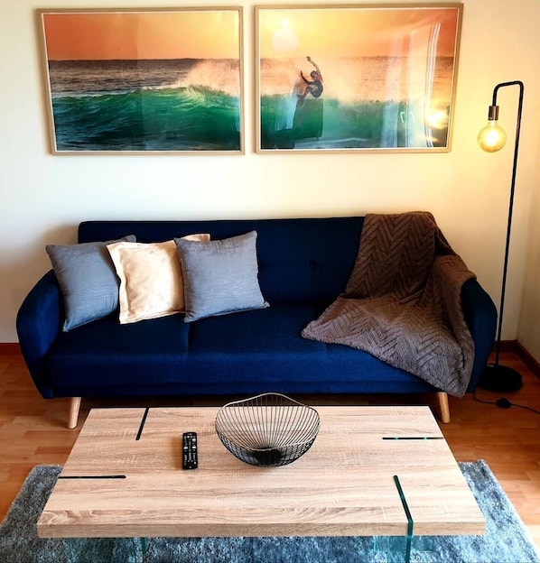 A living room with a dark blue sofa, two cushions, a throw blanket, a wooden coffee table with a wire bowl, a remote control, and two framed artworks depicting ocean scenes.