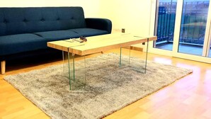Ein Zimmer mit einem dunkelblauen Sofa, einem Couchtisch aus Holz mit Glasbeinen und einem grauen, flauschigen Teppich auf Holzdielen.
