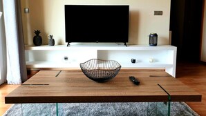 Ein modernes Wohnzimmer mit einem Holztisch, einem schwarzen Drahtkorb, einer Fernbedienung und einem Fernseher auf einer weißen Unterhaltungseinheit.