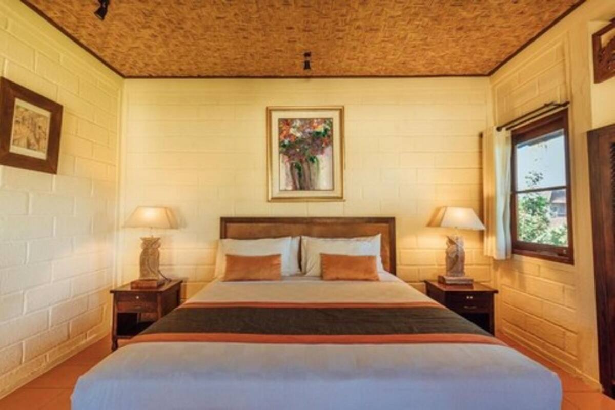 Un dormitorio con cabecera de madera, dos mesitas de noche con lámparas, una obra de arte enmarcada sobre la cama y una ventana a la derecha.
