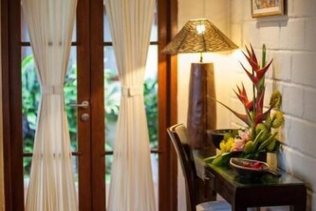 Una habitación con puerta de madera, cortinas blancas, una mesa con lámpara, sillas y una planta decorativa.