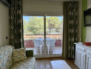 Habitació amb sofà, taula de menjador amb cadires, televisió penjada a la paret i balcó amb vistes a arbres i una tanca.