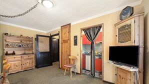 Een woonkamer met een televisie, houten kasten, een stoel en een zichtbare keuken door een open deur.