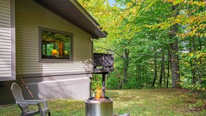 Un poêle en métal, posé sur une plateforme en bois à l’extérieur d’une maison, avec une chaise à proximité.