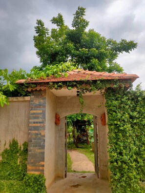 A garden archway with a tiled roof, surrounded by lush greenery and a pathway leading through it.