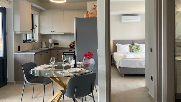 A modern kitchen with white cabinets, a dining area with a glass table and chairs, and a bedroom with a bed and a nightstand. - MOSSA CITY APARTMENTS (Chania)