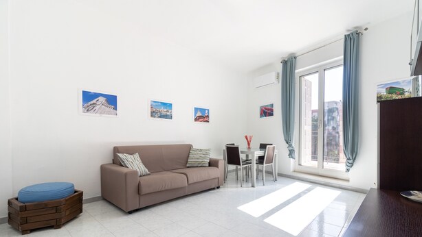 A modern living room with a sofa, a dining table with chairs, a small wooden stool, and a large window with curtains. - Esperia Home penthouse in Bari with private terrace, Wi-Fi, and air conditioning (Bari)