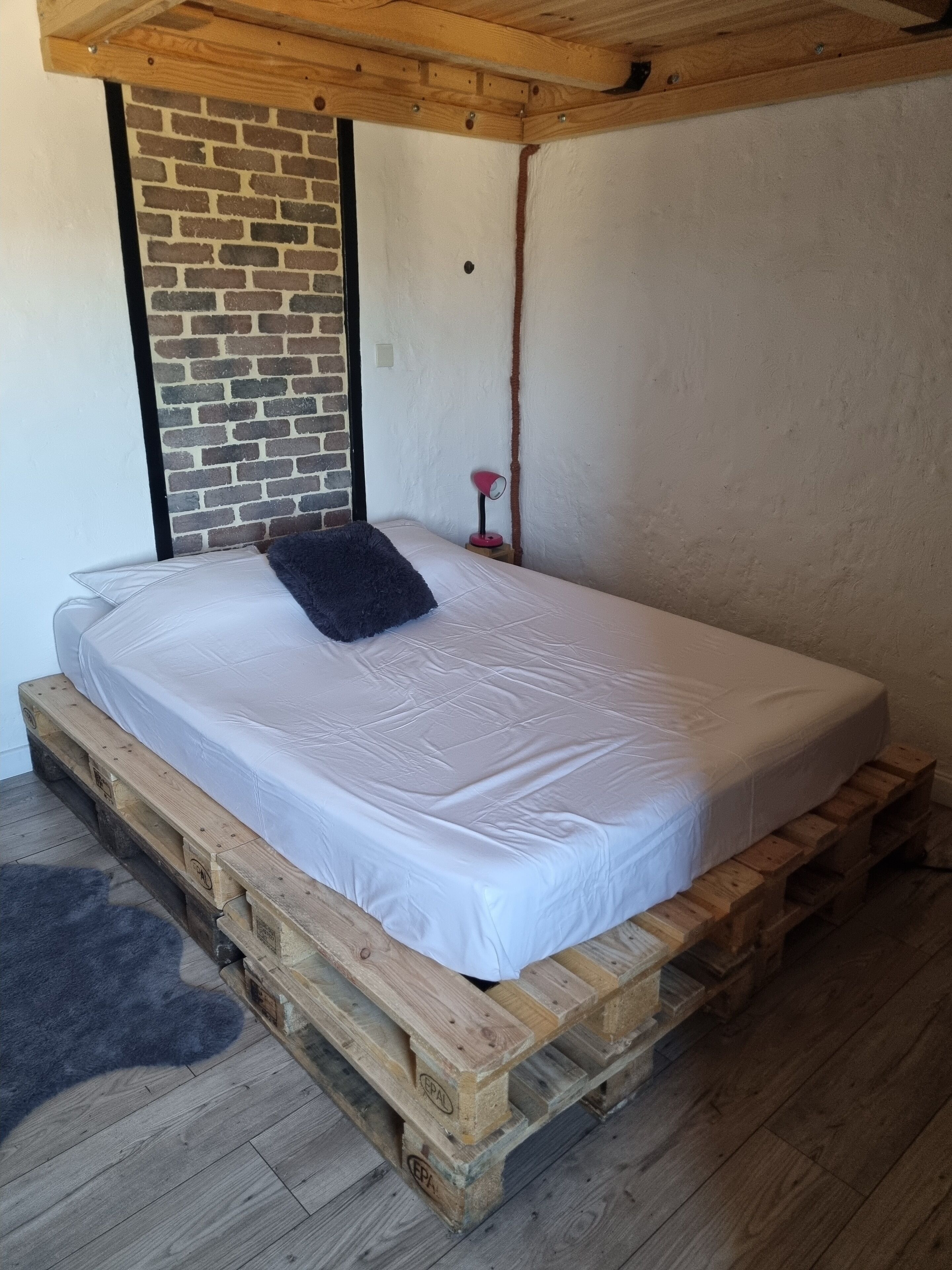 A single bed on a wooden pallet base with a white mattress and a dark pillow. A brick wall and a wooden beam ceiling are visible.