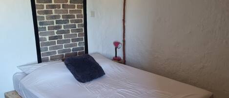 A single bed on a wooden pallet base with a white mattress and a dark pillow. A brick wall and a wooden beam ceiling are visible.