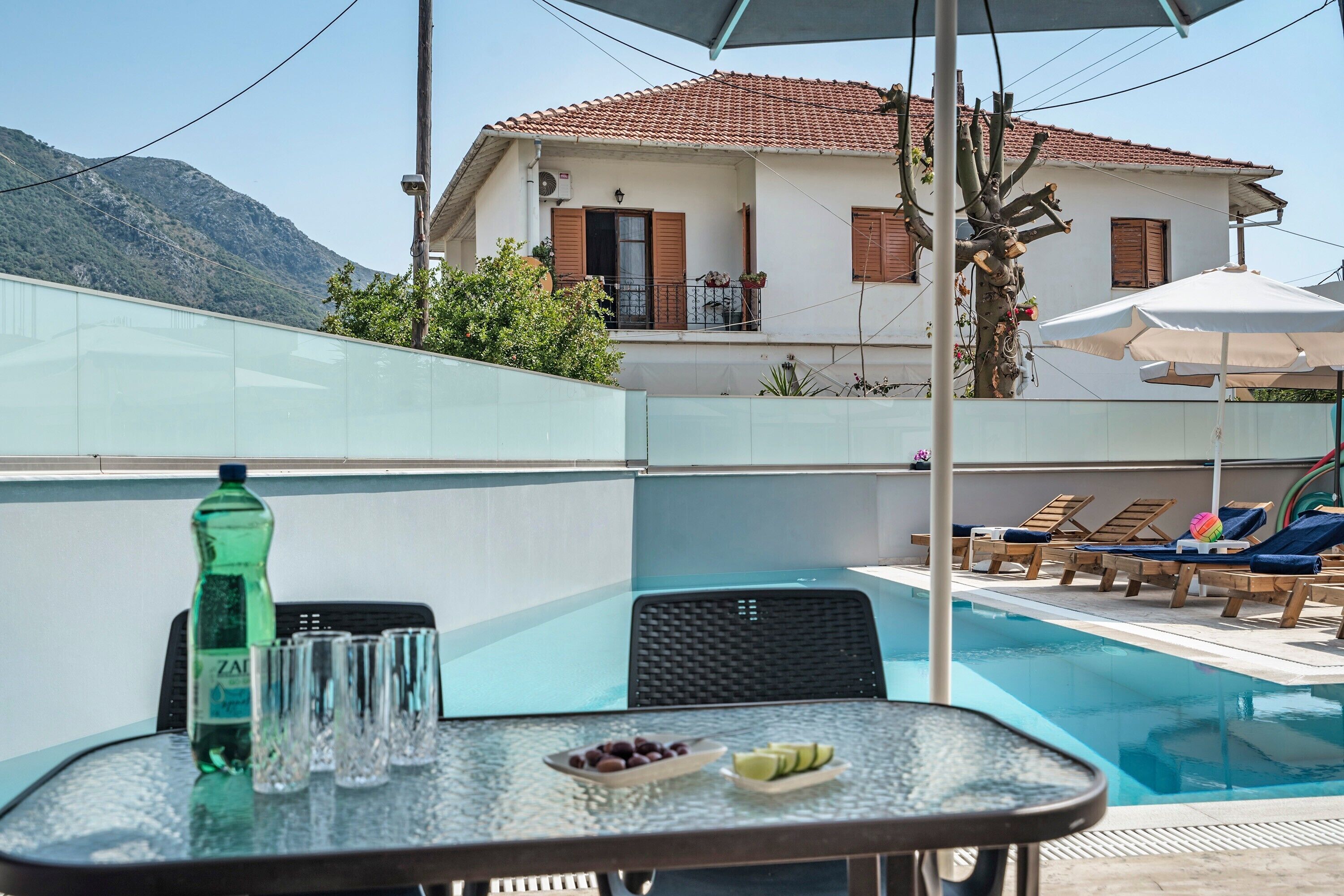 A poolside area with lounge chairs, a table with a bottle and glasses, and a building with a tiled roof in the background.